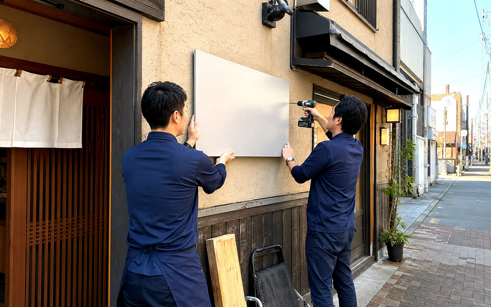 店舗入口で2人で中型看板を電動ドリルで取付けているスタッフ